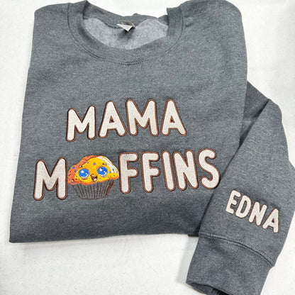 A folded heather gray sweatshirt on a white background, featuring the text 'MAMA MOFFINS' in raised cream letters with brown outlines, where the letter O is replaced by a colorful cupcake graphic. The right sleeve has the name 'EDNA' embroidered in matching cream letters.