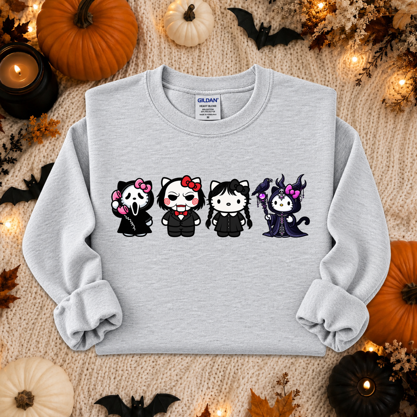 A light gray sweatshirt laid flat on a cozy beige knit blanket, surrounded by Halloween decorations including pumpkins, candles, leaves, and bats. The sweatshirt features a graphic of four cartoon hello kitty characters dressed in Halloween costumes, styled in a warm autumn flat-lay setup.