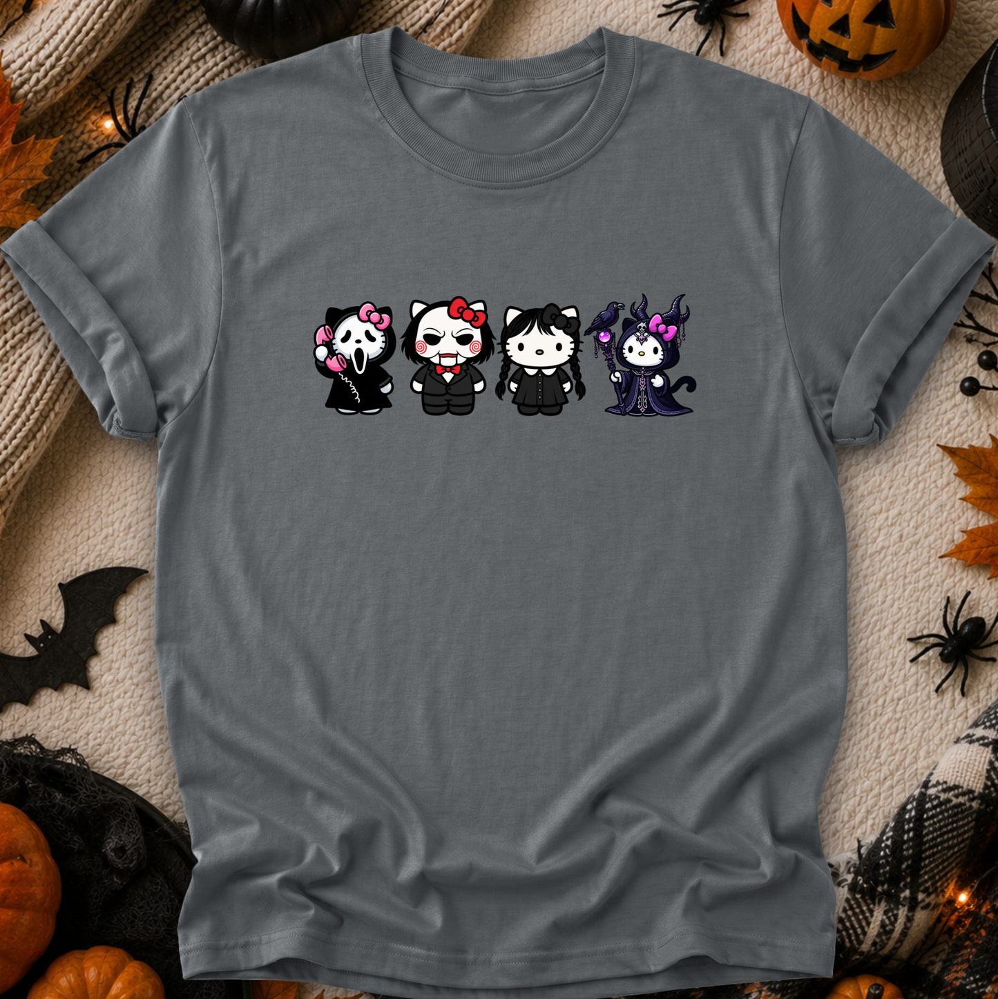 A dark gray short-sleeve t-shirt laid flat on a cozy beige knit blanket surrounded by Halloween decorations, including pumpkins, a spider, a bat, and spiderwebs. The shirt features a graphic print of four cute, chibi-style vampire characters in black outfits with pink, red, and purple accents.