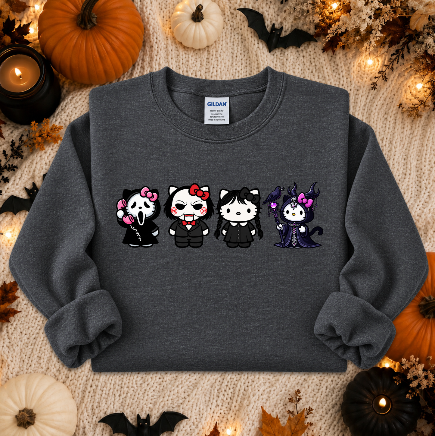 A dark gray crewneck sweatshirt featuring a graphic of four cute cartoon cats dressed as Halloween characters, laid flat on a cozy beige knit blanket surrounded by autumn decorations like pumpkins, bats, and candles. A Gildan brand tag is visible inside the collar.