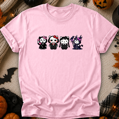 A light pink short-sleeve t-shirt with a graphic of four cute, chibi-style Halloween characters in black outfits, laid flat on a cozy beige fabric background decorated with autumn and Halloween props like pumpkins, a spider, and a bat.