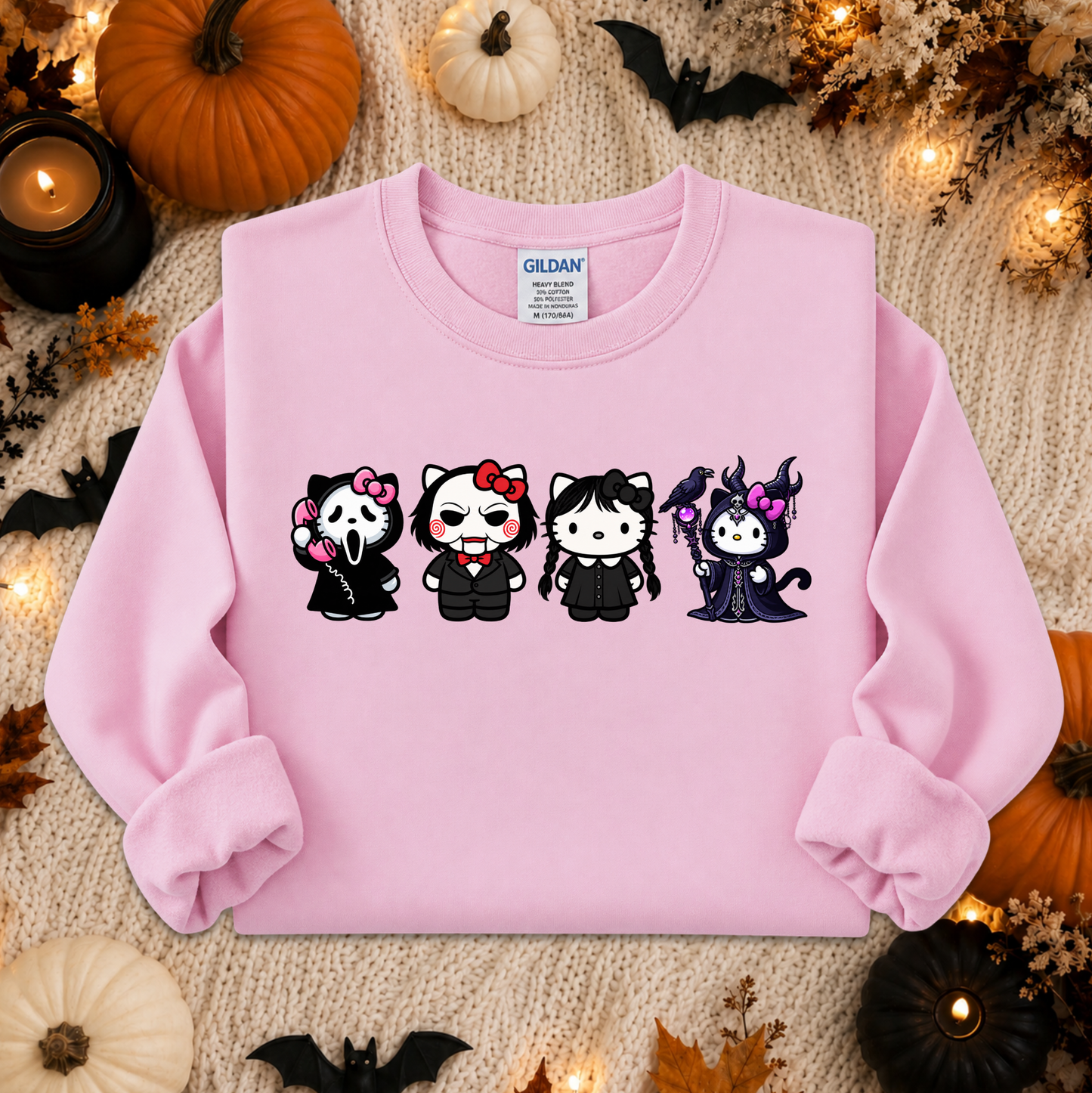 A light pink crewneck sweatshirt featuring a graphic of four cartoon cats in Halloween costumes, styled in a flat lay with autumn decorations like pumpkins, candles, and leaves. The brand 'Gildan' is visible on the inner neck label.