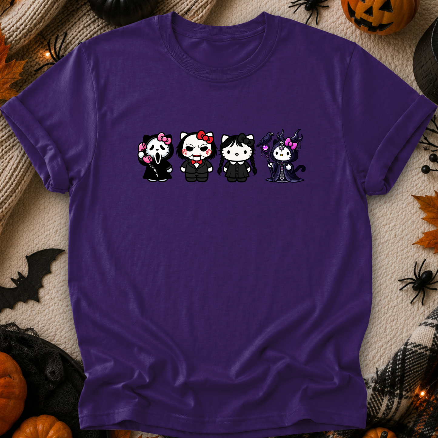 A purple short-sleeve t-shirt laid flat on a textured beige fabric background, surrounded by Halloween decorations including pumpkins, spiders, and a bat. The shirt features a graphic of four cartoon characters in black outfits with pink, white, and black details.