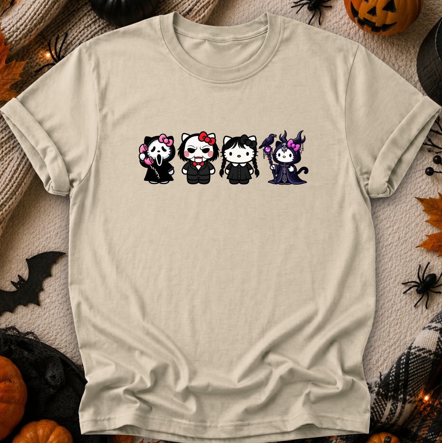 A beige graphic t-shirt featuring four cute, chibi-style cartoon cats in Halloween costumes, laid flat on a cozy beige fabric background surrounded by Halloween decorations like pumpkins, a spider, and a bat.