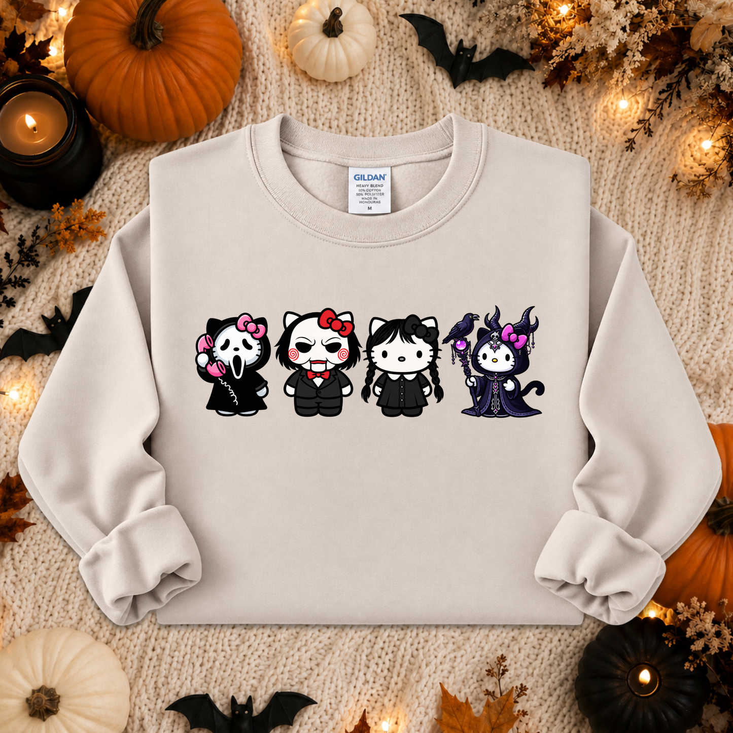 A light beige crewneck sweatshirt featuring a graphic of four cute, stylized Hello Kitty characters dressed in Halloween costumes, presented in a flat-lay arrangement with autumn and Halloween decorations.