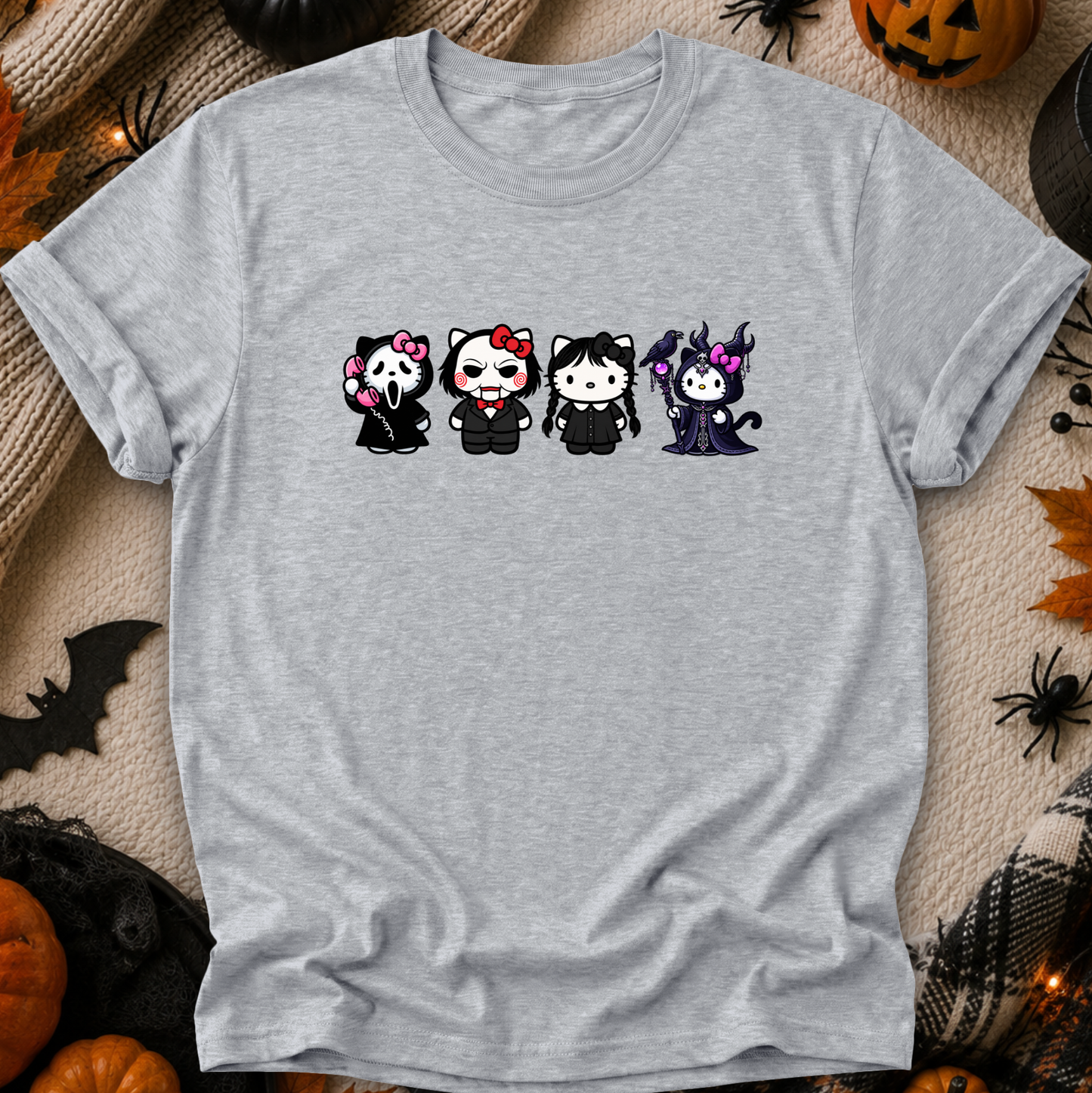 A light heather gray short-sleeve t-shirt laid flat on a cozy beige fabric background with Halloween decorations around it. The shirt features a graphic of four cute cartoon cats in black outfits with pink, red, and purple accents.