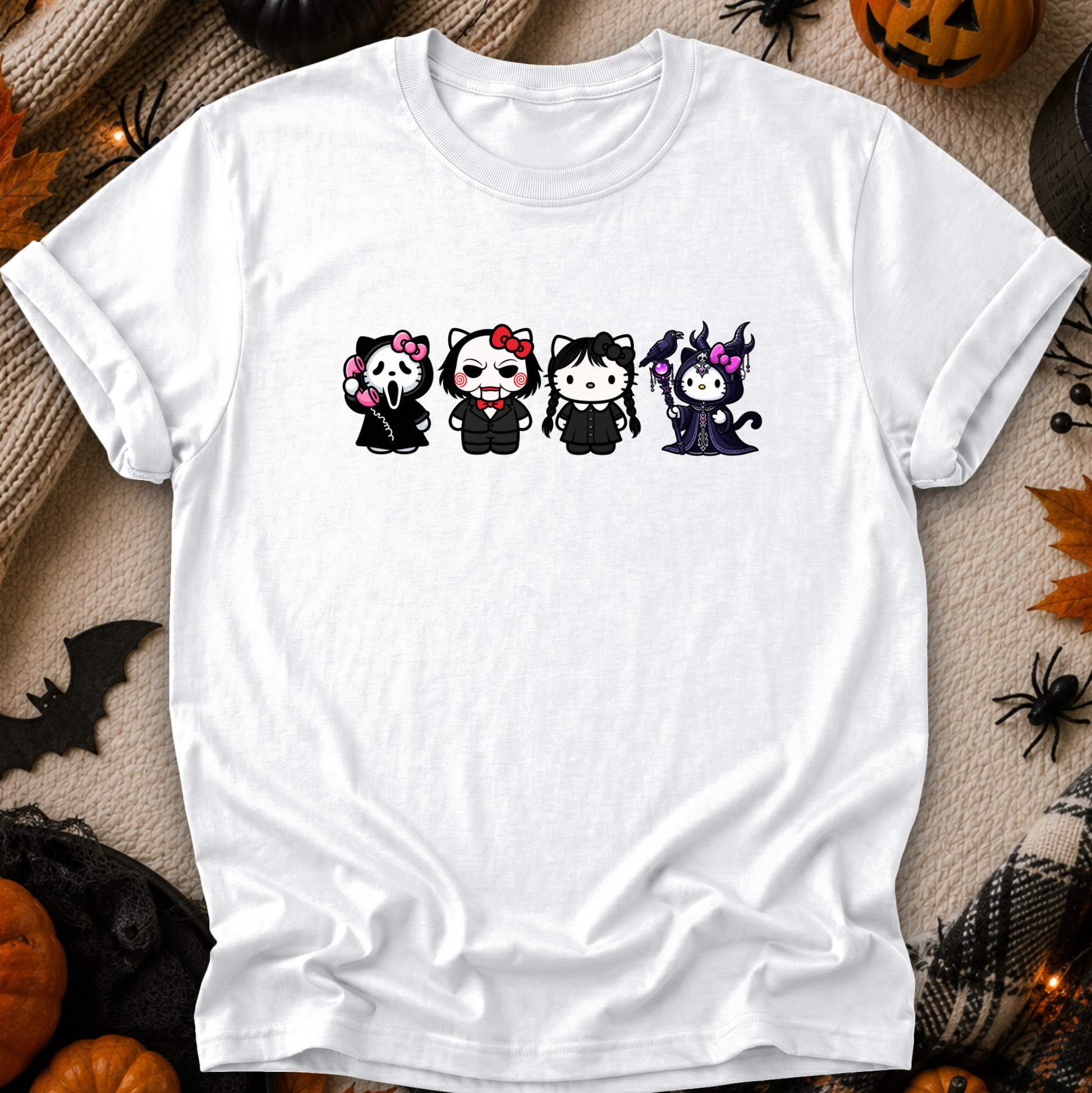 A white short-sleeve crewneck t-shirt laid flat on a cozy beige fabric background with Halloween decorations around it. The shirt features a graphic of four cute cartoon cat characters dressed in black gothic outfits, including a vampire, a skeleton, a cat in a red bow, and a witch.