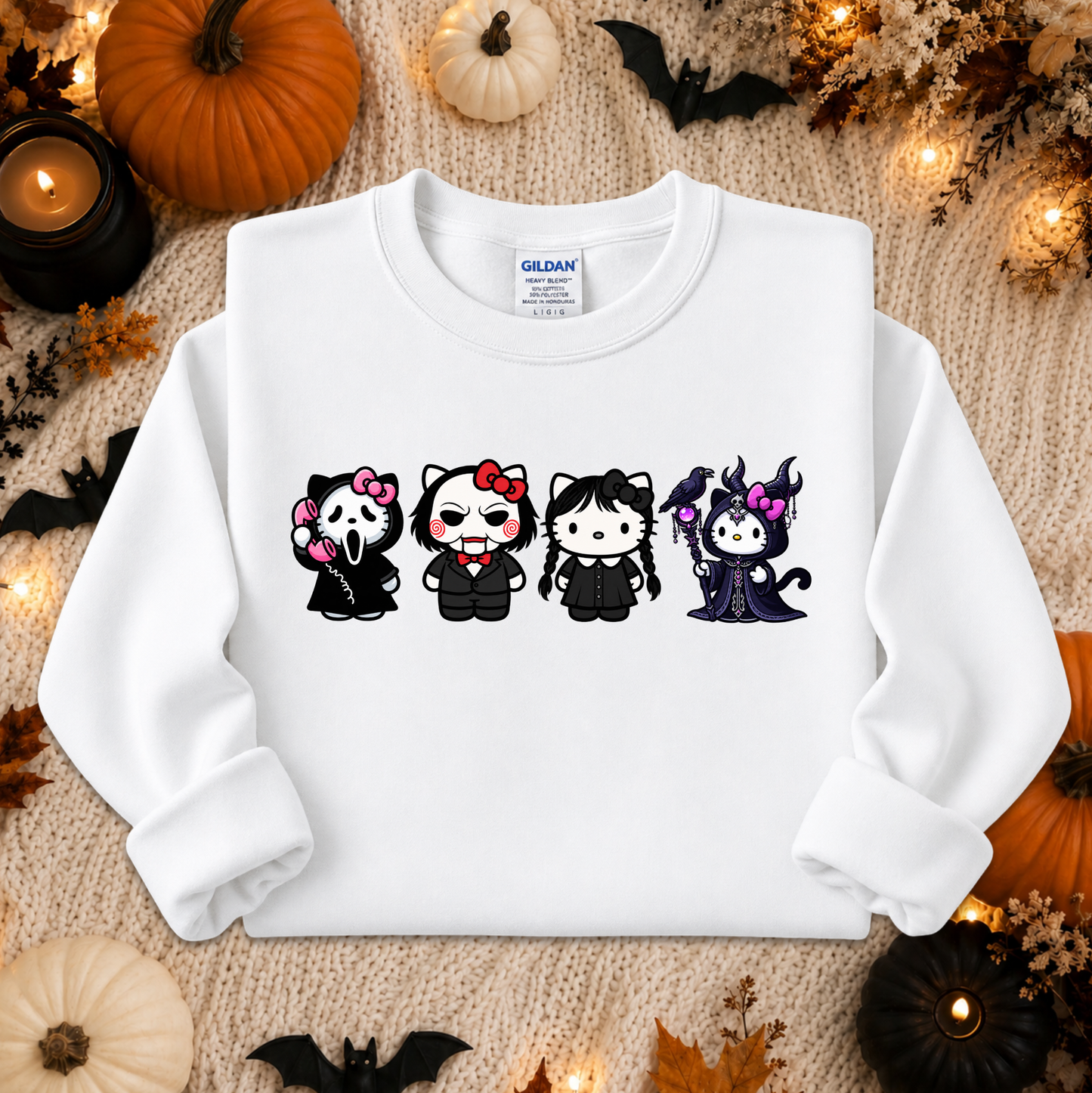 A white long-sleeve crewneck sweatshirt laid flat on a textured beige knit blanket, surrounded by Halloween decorations including pumpkins, candles, leaves, and bats. The sweatshirt features a graphic of four cute cartoon characters in black outfits with pink and purple accents.