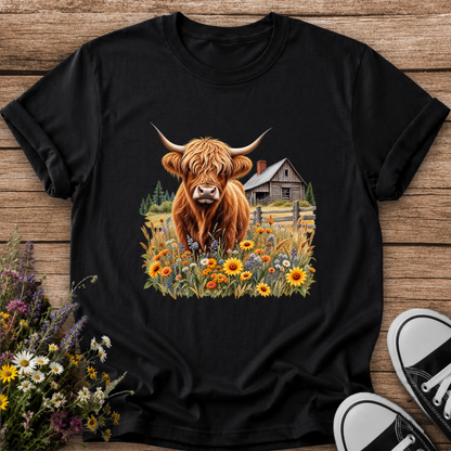 black tshirt with a graphic of a highland cow standing in a field of wildflowers in front of a barn