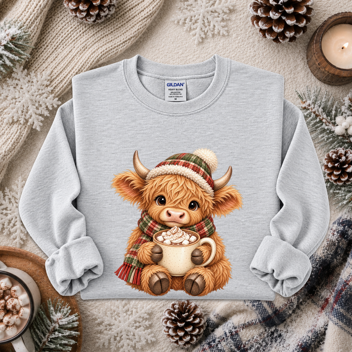 A light gray long-sleeve sweatshirt laid flat, featuring a cute cartoon Highland cow wearing a plaid hat and scarf while holding a mug of hot cocoa. The sweatshirt is styled in a cozy winter flat-lay with pinecones, a candle, knit fabric, and snowflake decorations.