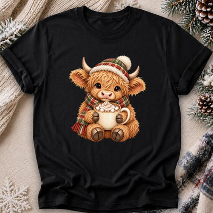 A black short-sleeve crewneck t-shirt featuring a cute cartoon Highland cow illustration wearing a red, green, and white plaid hat and scarf while holding a mug of hot chocolate. The t-shirt is displayed in a flat-lay setting on a cozy beige blanket with knitted sweaters, pinecones, and evergreen branches.