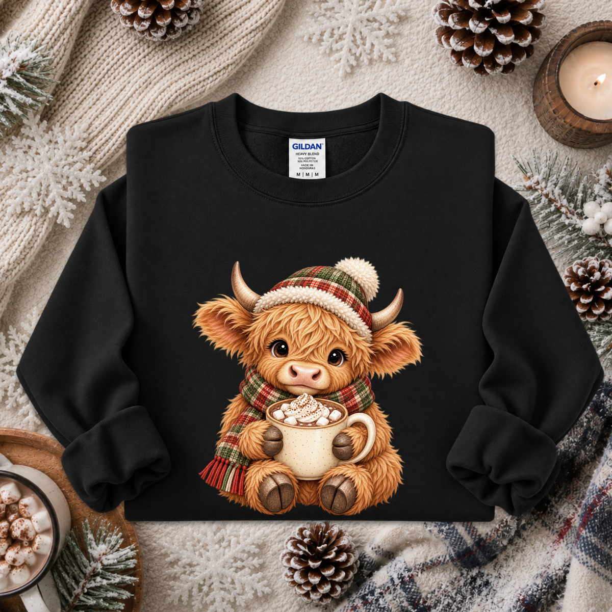 A black crewneck sweatshirt featuring a cute cartoon illustration of a Highland cow in a plaid hat and scarf holding a mug of hot cocoa, styled in a cozy flat-lay with pinecones, a knitted blanket, and a candle.