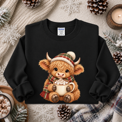 A black crewneck sweatshirt featuring a cute cartoon illustration of a Highland cow in a plaid hat and scarf holding a mug of hot cocoa, styled in a cozy flat-lay with pinecones, a knitted blanket, and a candle.