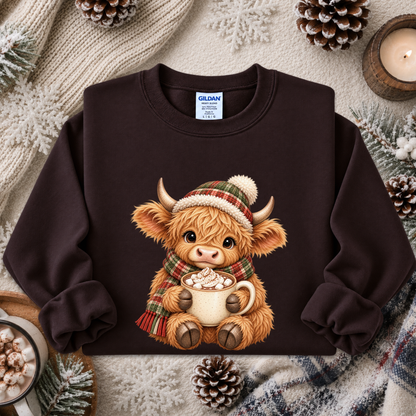 A dark brown crewneck sweatshirt featuring a cute graphic of a Highland cow wearing a green and red plaid hat and scarf while holding a mug of hot chocolate. The sweatshirt is styled in a cozy flat-lay arrangement with a knitted cream sweater, pinecones, a candle, and a plaid blanket.