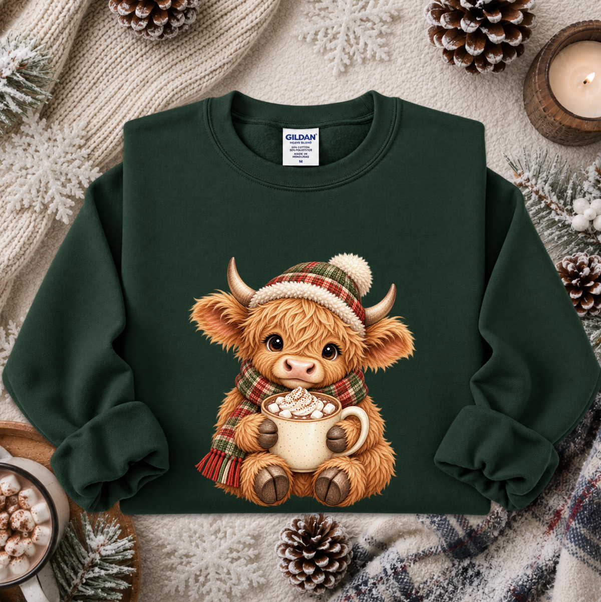 A dark green crewneck sweatshirt featuring a cute graphic of a fluffy Highland cow wearing a plaid hat and scarf while holding a mug of hot chocolate. The sweatshirt is styled in a cozy flat-lay arrangement with a knit sweater, pinecones, and a candle.