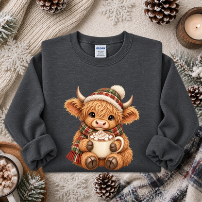A dark gray crewneck sweatshirt featuring a cute cartoon Highland cow graphic wearing a plaid hat and scarf while holding a mug of hot chocolate. The sweatshirt is styled in a cozy winter flat-lay with a knit blanket, pinecones, and a candle.