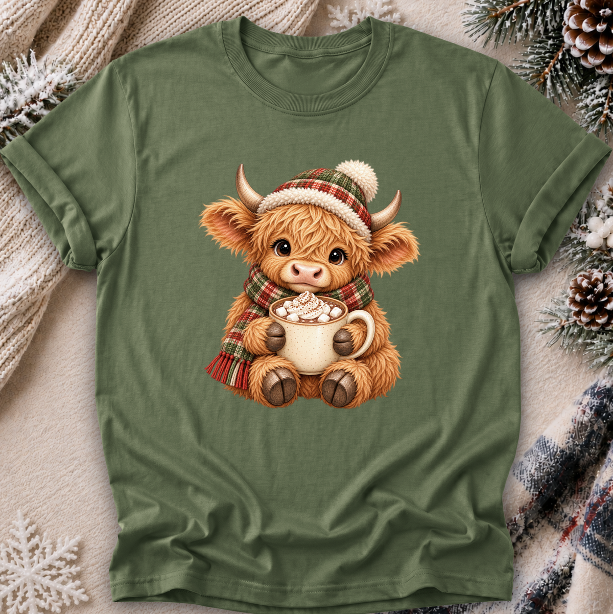 An olive green short-sleeve t-shirt featuring a cute cartoon Highland cow graphic wearing a plaid hat and scarf while holding a mug of hot cocoa. The t-shirt is displayed in a cozy flat-lay setting with a cream knit blanket, pinecones, and a plaid scarf.