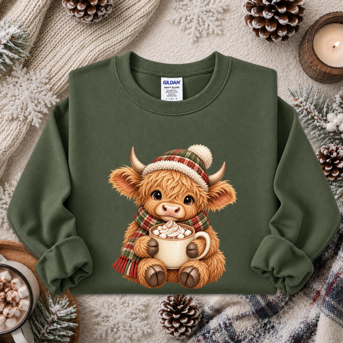An olive green crewneck sweatshirt featuring a cute cartoon Highland cow graphic wearing a plaid hat and scarf while holding a mug of hot cocoa. The sweatshirt is styled in a cozy flat-lay arrangement with a knitted blanket, pinecones, and a lit candle.