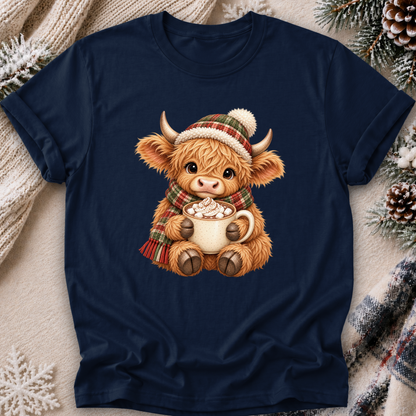A navy blue short-sleeve t-shirt with a central graphic of a cute Highland cow wearing a red and green plaid hat and scarf, holding a mug of hot cocoa. The t-shirt is displayed in a flat-lay setting with a cozy knit blanket, pinecones, and snowflakes.