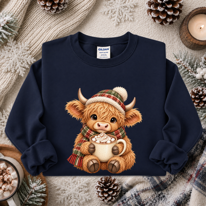 A navy blue crewneck sweatshirt featuring a cute graphic of a fluffy Highland cow wearing a green and red plaid hat and scarf while holding a mug of hot cocoa. The sweatshirt is styled in a cozy flat-lay arrangement with a knitted cream sweater, pinecones, a candle, and winter decorations.