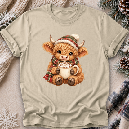 A beige crewneck t-shirt featuring a cute cartoon Highland cow graphic wearing a red and green plaid knit hat and matching scarf while holding a mug of hot cocoa with marshmallows. The t-shirt is displayed flat on a cozy background with pinecones, a knit sweater, and a plaid blanket.