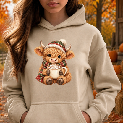 A woman wearing a beige hoodie with a graphic of a fluffy Highland cow wearing a plaid hat and scarf while holding a mug of hot chocolate. The background shows an autumn outdoor scene with pumpkins and orange foliage.