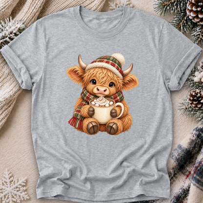 A light heather gray short-sleeve t-shirt featuring a cute graphic of a fluffy brown Scottish highland cow wearing a red and green plaid knit hat and matching scarf, holding a mug of hot cocoa with marshmallows. The t-shirt is styled in a cozy flat-lay setting with a cream knit sweater, pinecones, and a plaid scarf.