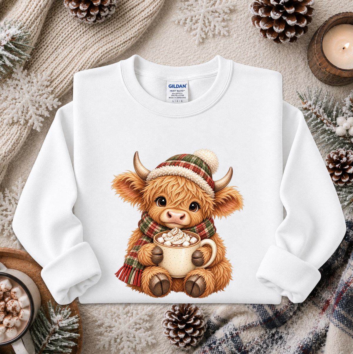 A white long-sleeve crewneck sweatshirt featuring a cute cartoon Highland cow wearing a red and green plaid hat and matching scarf while holding a mug of hot cocoa. The sweatshirt is displayed in a cozy flat-lay setting with pinecones, a candle, and knitted blankets.