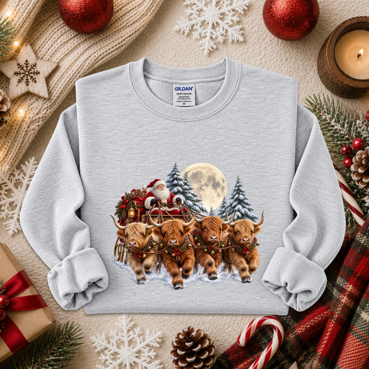 A light gray crewneck sweatshirt featuring a festive Christmas graphic of four Highland cows pulling a sleigh with Santa and a full moon in the background. The sweatshirt is styled in a cozy holiday flat-lay with a knitted sweater, red ornaments, pinecones, candy canes, and a plaid scarf.