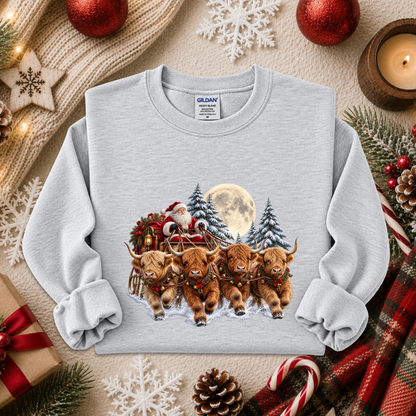 A light gray crewneck sweatshirt featuring a festive Christmas graphic of four Highland cows pulling a sleigh with Santa and a full moon in the background. The sweatshirt is styled in a cozy holiday flat-lay with a knitted sweater, red ornaments, pinecones, candy canes, and a plaid scarf.