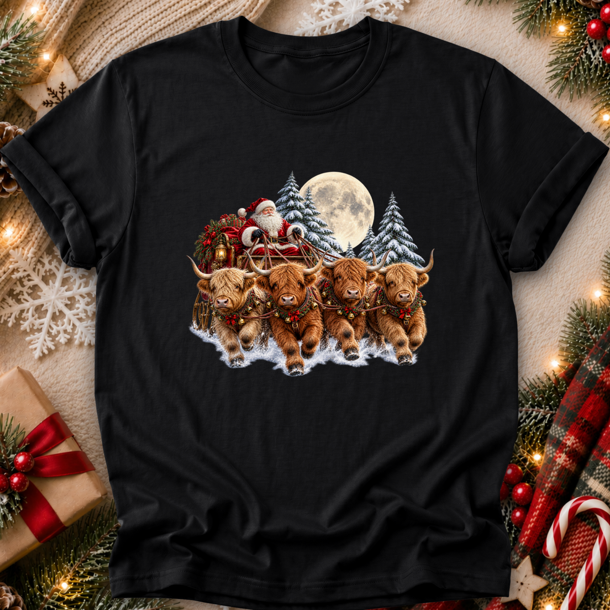 A black short-sleeve crewneck t-shirt featuring a festive Christmas graphic of four Highland cows pulling Santa's sleigh through the snow, surrounded by pine trees, a full moon, and a cozy holiday flat-lay background with lights, gifts, and plaid fabric.