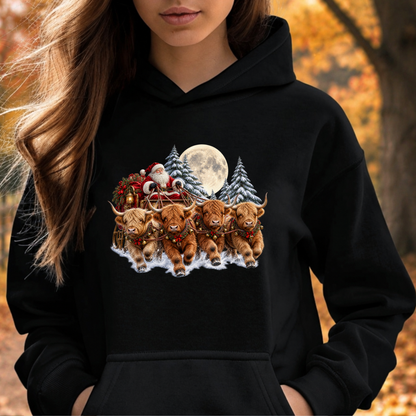 A woman wearing a black hoodie with a colorful graphic print of Santa Claus, a full moon, and four Highland cattle in a snowy landscape, set against an autumn outdoor background.