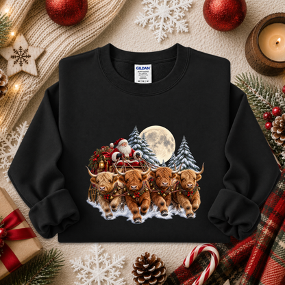 A black crewneck sweatshirt featuring a festive Christmas graphic of four Highland cows pulling Santa's sleigh in the snow, surrounded by pine trees and a full moon. The sweatshirt is displayed in a cozy holiday flat-lay setting with red ornaments, a candle, a gift box, and a plaid scarf.