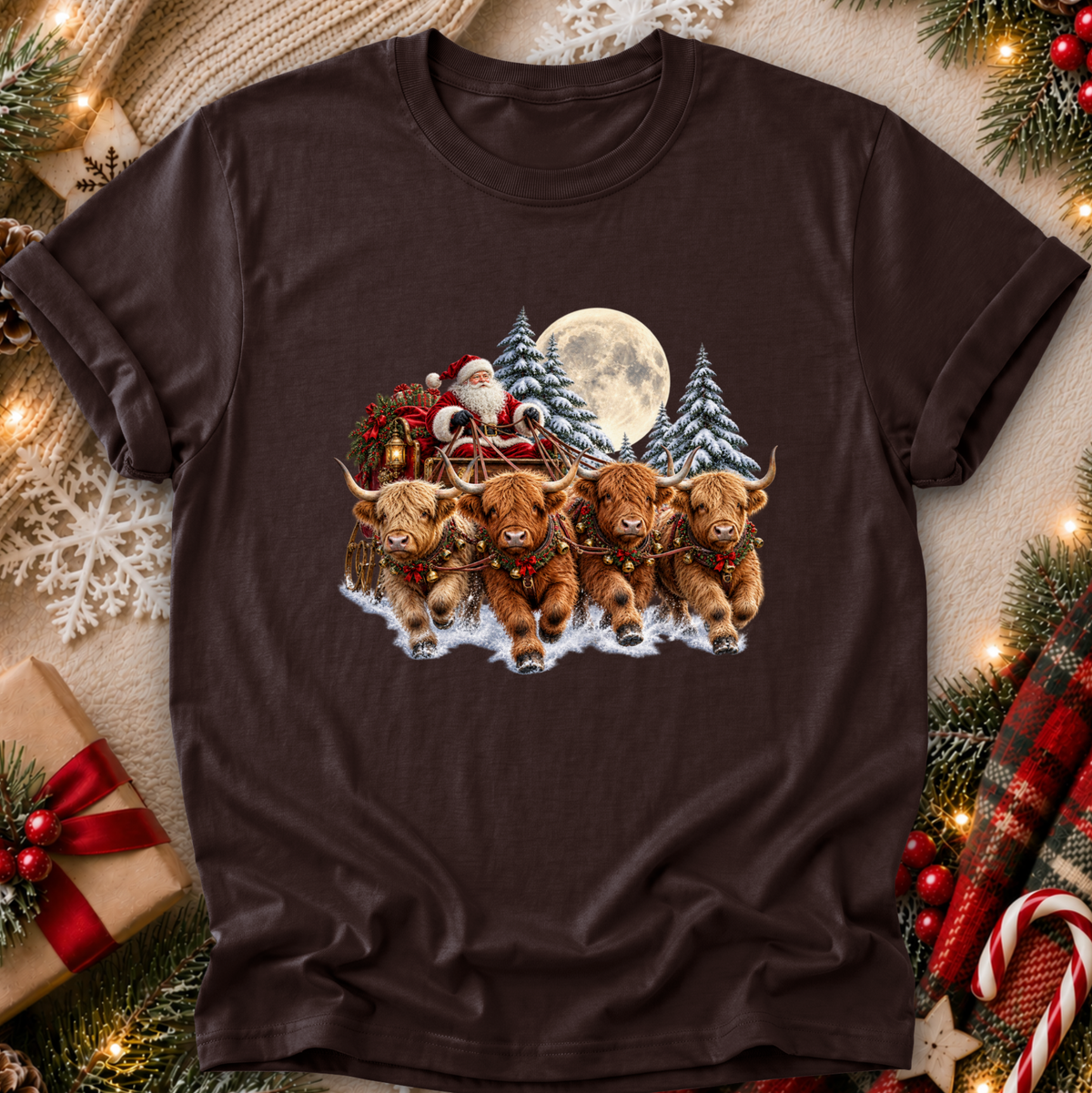 A dark brown short-sleeve crewneck t-shirt featuring a festive Christmas graphic of four Highland cows pulling Santa's sleigh through the snow, surrounded by pine trees, a full moon, and Santa Claus. The t-shirt is displayed in a styled flat-lay setting with holiday decorations like pine branches, ornaments, a wrapped gift, and a plaid blanket.