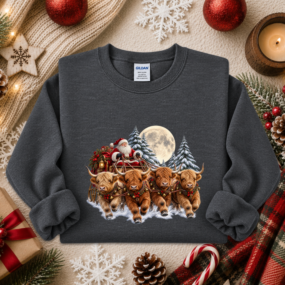 A dark gray crewneck sweatshirt featuring a festive graphic of four Highland cows pulling a sleigh with Santa Claus under a full moon, presented in a cozy holiday flat-lay with knitted scarves, ornaments, and candy canes.