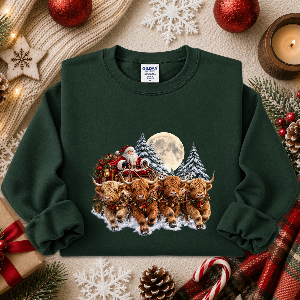 A dark green crewneck sweatshirt featuring a festive Christmas graphic of four Highland cows pulling Santa's sleigh in the snow, surrounded by holiday decorations including a candle, pinecones, candy canes, and red ornaments on a beige knit blanket.