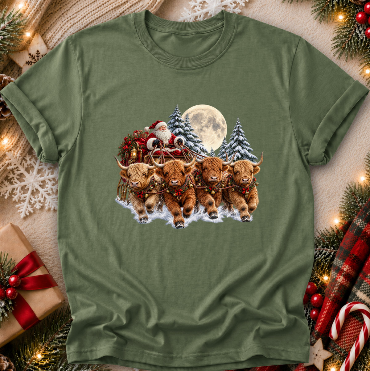 An olive green short-sleeve t-shirt with a festive Christmas graphic of four Highland cows pulling Santa's sleigh under a full moon, presented in a cozy holiday flat-lay setting with pine branches, ornaments, and a gift box.