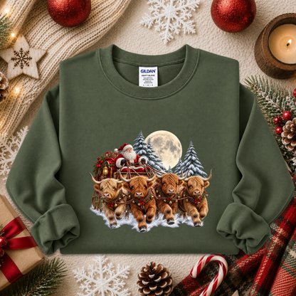 An olive green crewneck sweatshirt by Gildan, featuring a festive Christmas graphic of four Highland cows pulling a sleigh with Santa Claus. The sweatshirt is styled in a cozy holiday flat-lay arrangement with red ornaments, a candle, pinecones, candy canes, and a knitted scarf.