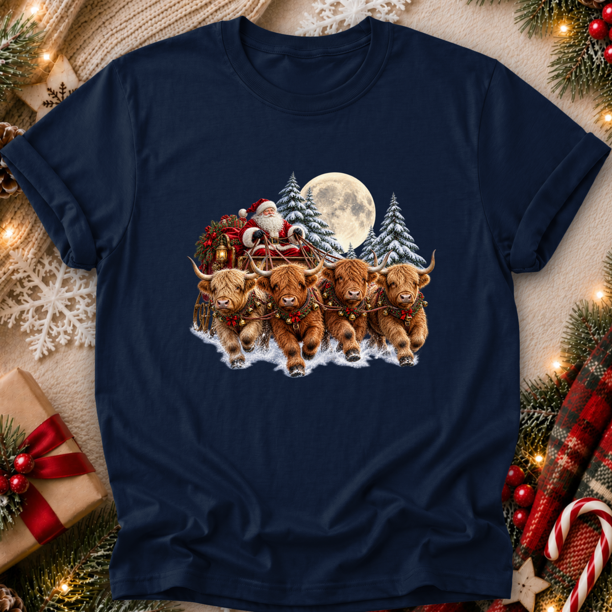 A navy blue short-sleeve t-shirt with a festive Christmas graphic of four Highland cows pulling Santa's sleigh under a full moon, displayed in a cozy holiday flat-lay setting with pine branches, lights, and a gift box.