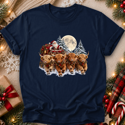 A navy blue short-sleeve t-shirt with a festive Christmas graphic of four Highland cows pulling Santa's sleigh under a full moon, displayed in a cozy holiday flat-lay setting with pine branches, lights, and a gift box.