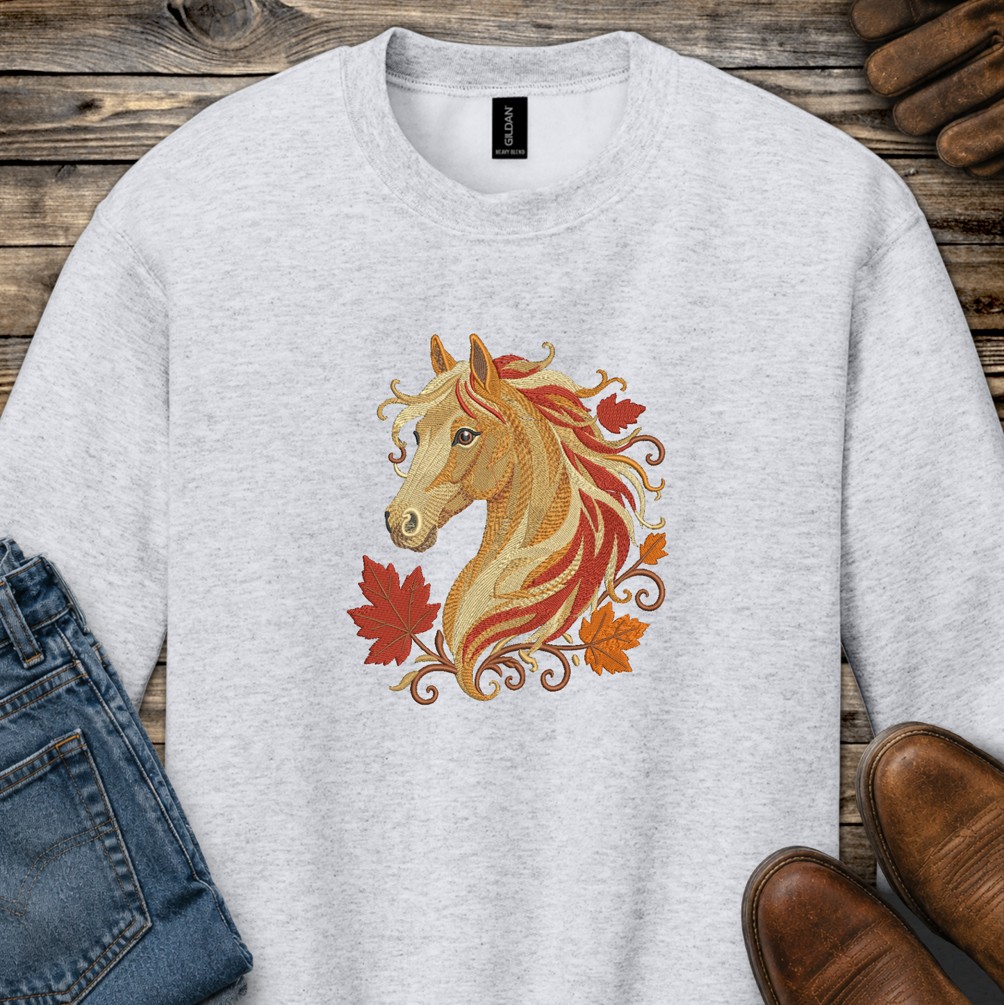 A light heather gray crewneck sweatshirt featuring a graphic of a golden horse head with red and orange autumn leaves. The sweatshirt is styled in a flat-lay arrangement on a rustic wooden background, accompanied by blue jeans and brown leather shoes.