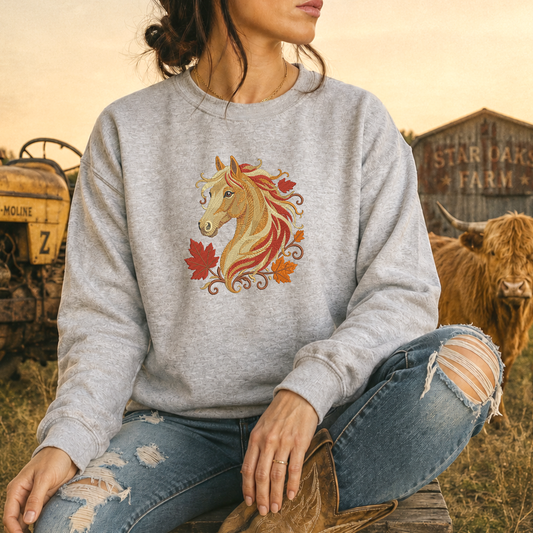 A woman wearing a light heather gray crewneck sweatshirt with a graphic of a golden horse head surrounded by red and orange autumn leaves. She is sitting outdoors in a rustic field with a cow and a barn in the background.