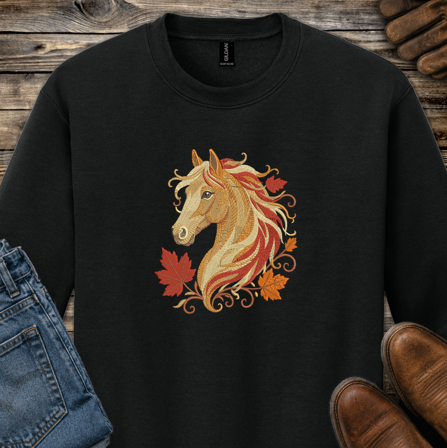 A black crewneck sweatshirt featuring a detailed graphic of a horse's head with flowing red, orange, and gold hair, surrounded by autumn maple leaves. The sweatshirt is styled in a flat-lay arrangement on a rustic wooden background with jeans, brown shoes, and leather boots.