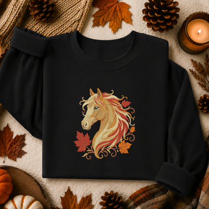 A black long-sleeve sweatshirt laid flat with rolled cuffs, featuring a decorative embroidered-style horse head graphic in tan, cream, red, and orange tones with autumn leaves around it. The sweatshirt is styled in a fall-themed flat lay with pinecones, leaves, a candle, and knit fabric.