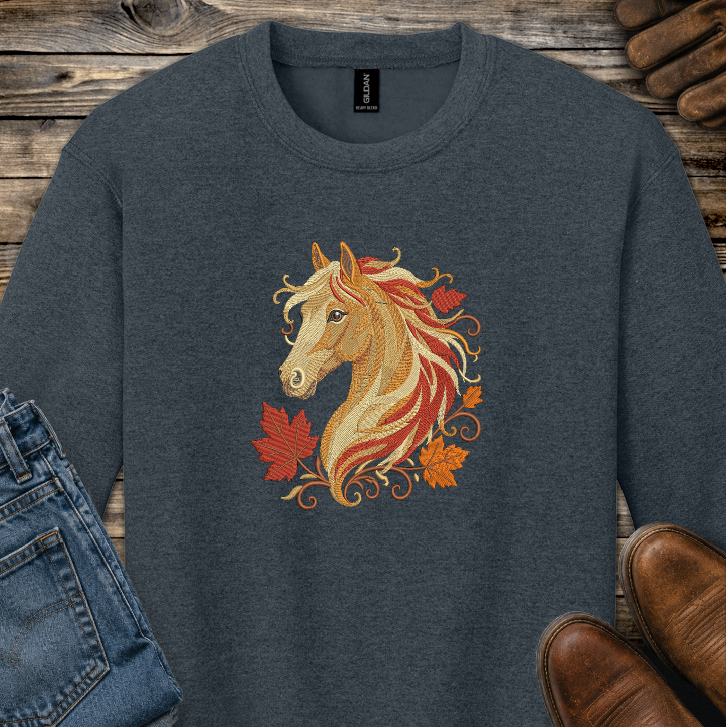 A dark heather gray crewneck sweatshirt featuring a graphic of a golden horse head surrounded by red and orange autumn leaves. The sweatshirt is styled in a flat-lay arrangement on a rustic wooden background, with blue jeans, brown leather shoes, and a pair of wooden shoes visible at the edges.