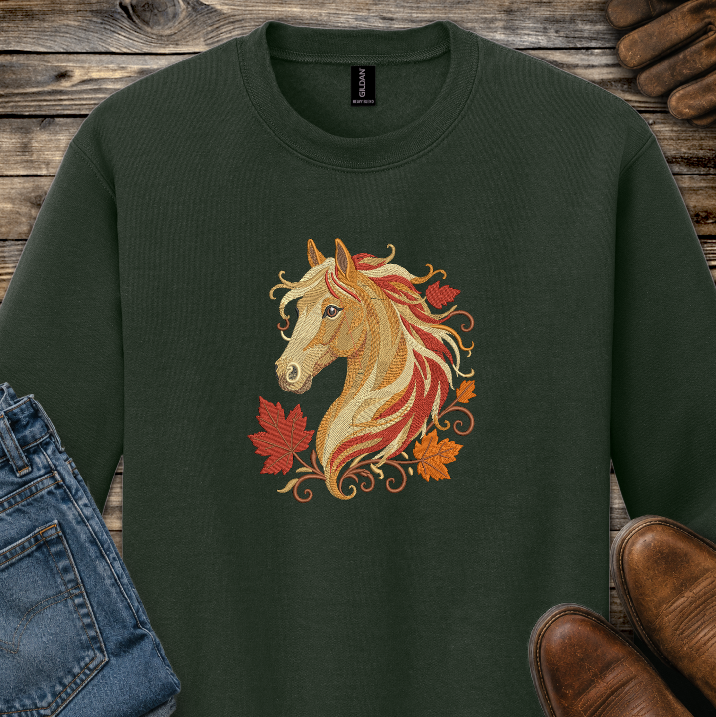 A dark forest green crewneck sweatshirt featuring a graphic of a horse head with flowing red, orange, and gold hair, surrounded by autumn maple leaves. The sweatshirt is styled in a flat-lay arrangement on a rustic wooden background, partially covered by blue jeans on the left and brown leather shoes on the right.