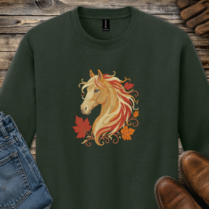 A dark forest green crewneck sweatshirt featuring a graphic of a horse head with flowing red, orange, and gold hair, surrounded by autumn maple leaves. The sweatshirt is styled in a flat-lay arrangement on a rustic wooden background, partially covered by blue jeans on the left and brown leather shoes on the right.