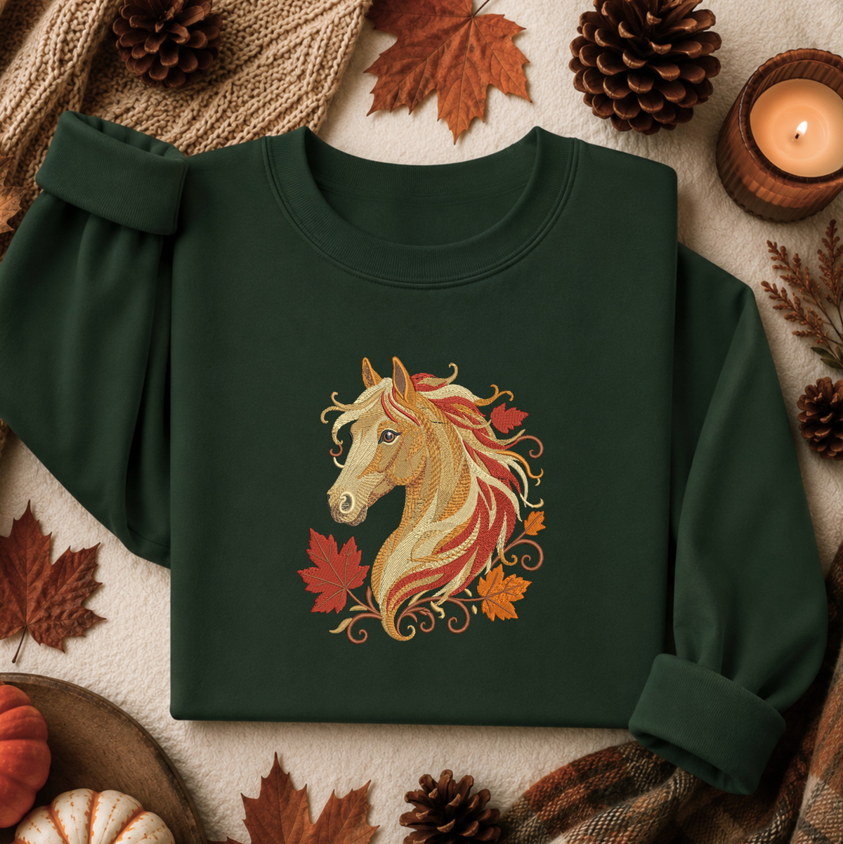 A dark forest green crewneck sweatshirt featuring a detailed graphic of a golden horse head with red and orange autumn leaves and swirls. The sweatshirt is styled in a cozy flat-lay arrangement with fall-themed props including pinecones, maple leaves, a lit candle, and a knit blanket.