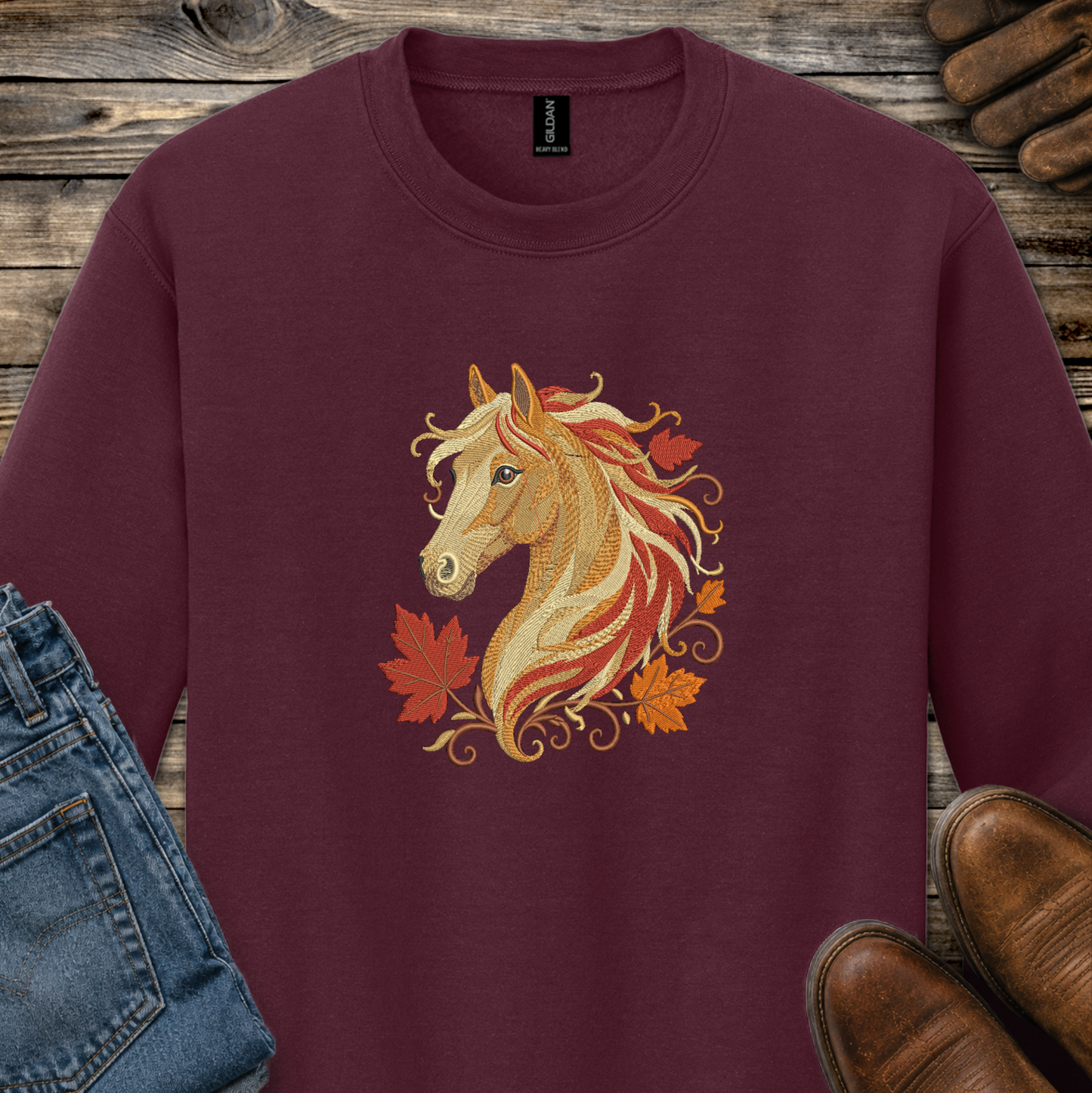 A maroon crewneck sweatshirt featuring a graphic of a golden horse head surrounded by orange and red autumn leaves. The sweatshirt is styled in a flat-lay arrangement on a rustic wooden background, partially covered by blue jeans, brown leather shoes, and a pair of brown boots.