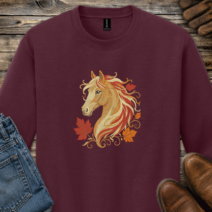 A maroon crewneck sweatshirt featuring a graphic of a golden horse head surrounded by orange and red autumn leaves. The sweatshirt is styled in a flat-lay arrangement on a rustic wooden background, partially covered by blue jeans, brown leather shoes, and a pair of brown boots.
